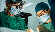 Navy surgeons in green scrubs and masks performing surgery; one surgeon wearing a headlamp with magnifying lenses, focusing on the procedure.