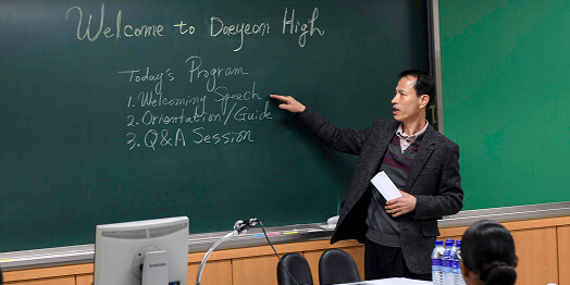 A high school teacher stands in front of a blackboard in a classroom, preparing to teach students.