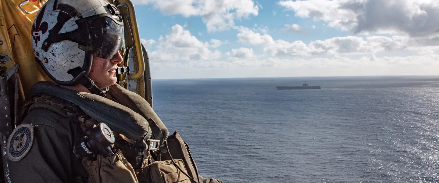A U.S. Navy Sailor looks out over the ocean from inside a helicopter as an aircraft carrier transits the sea.