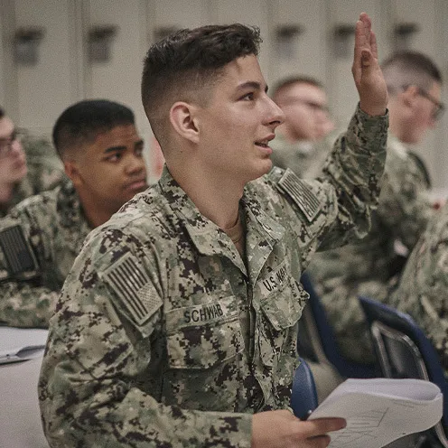 A student at Naval Nuclear Power School wearing his camouflage uniform raises a hand to answer a question while seated in a classroom with other Sailors.